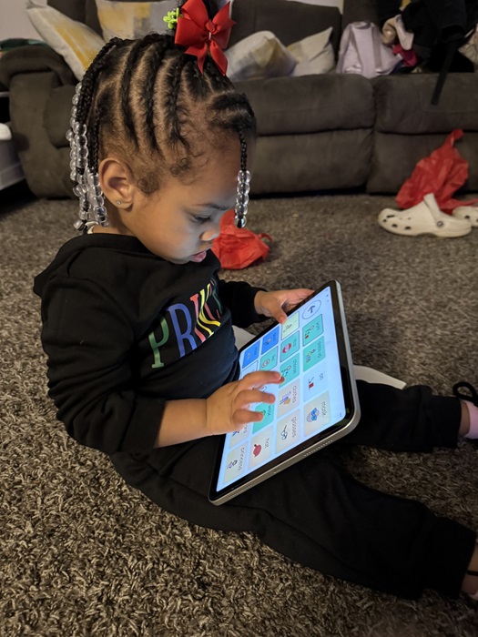 A young child sitting upright on the floor, using an AAC device that's in their lap