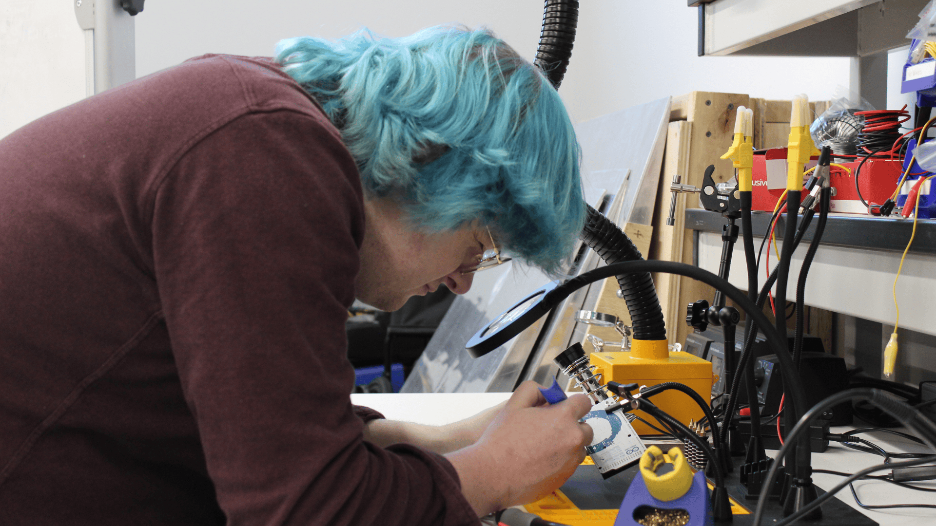 A man with blue hair does detailed electronics work under a magnifying glass at a laboratory workbench