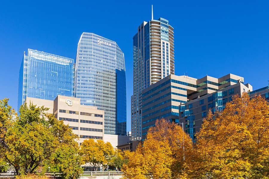 A view of downtown Denver high‑rise buildings alongside CU Denver buildings, with autumn trees in the foreground under a clear blue sky.